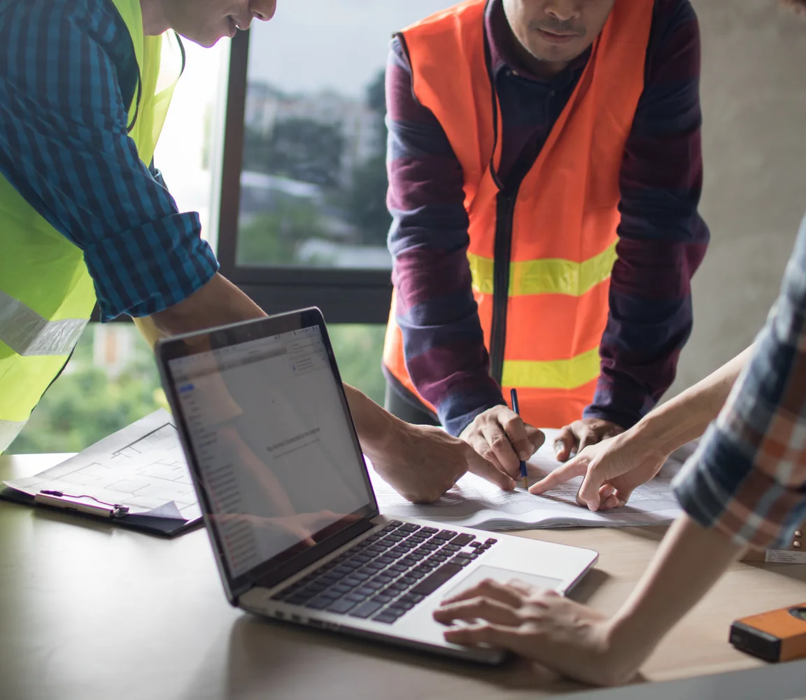 Three engineers in hard hats and high-visibility vests reviewing drawings around a laptop on site
