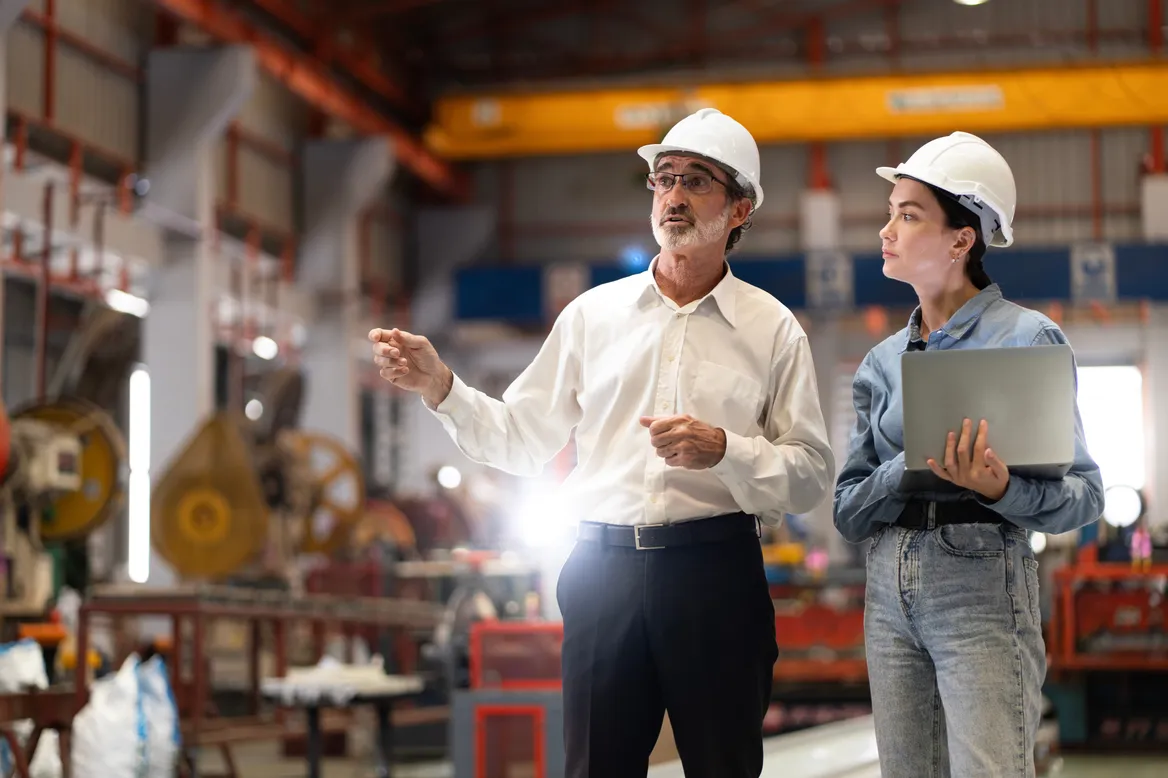 Two engineers in hard hats reviewing work on a laptop on an industrial workshop floor