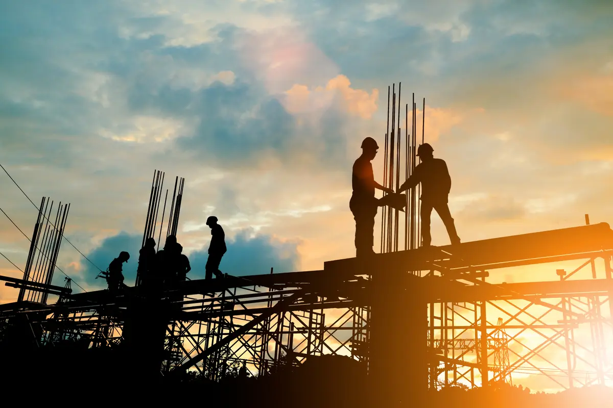 Silhouetted steelworkers on a reinforced concrete deck with protruding rebar cages at sunset