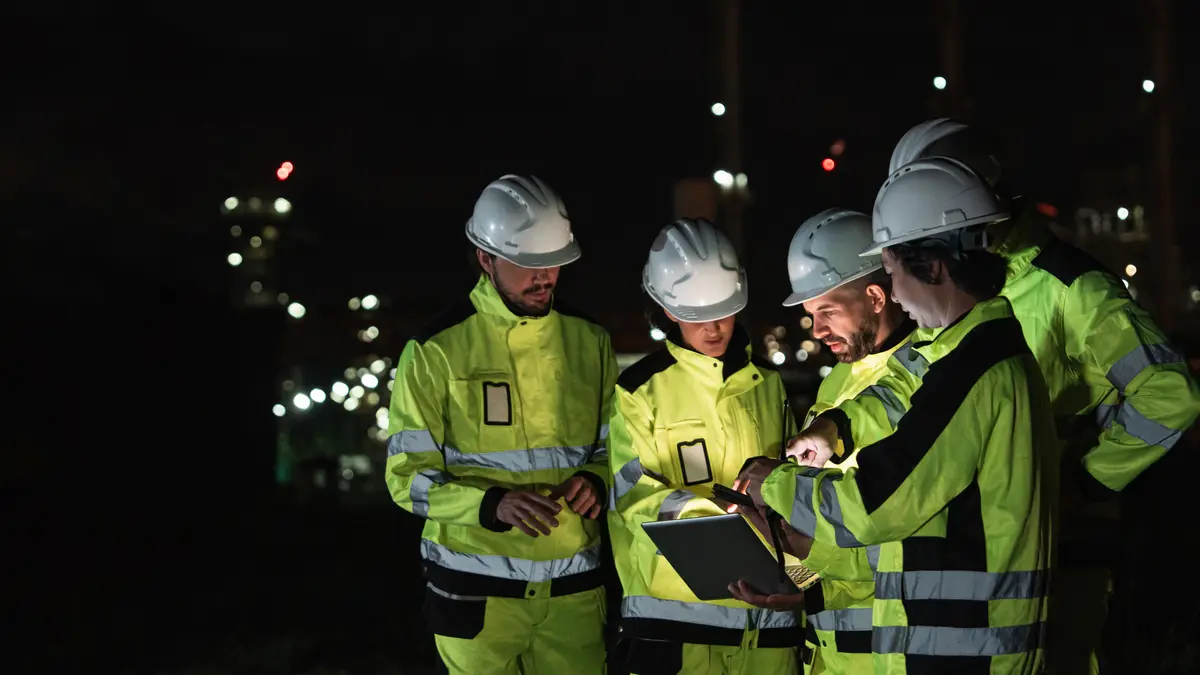 Engineers in hard hats reviewing a laptop at night, with a lit process plant behind them